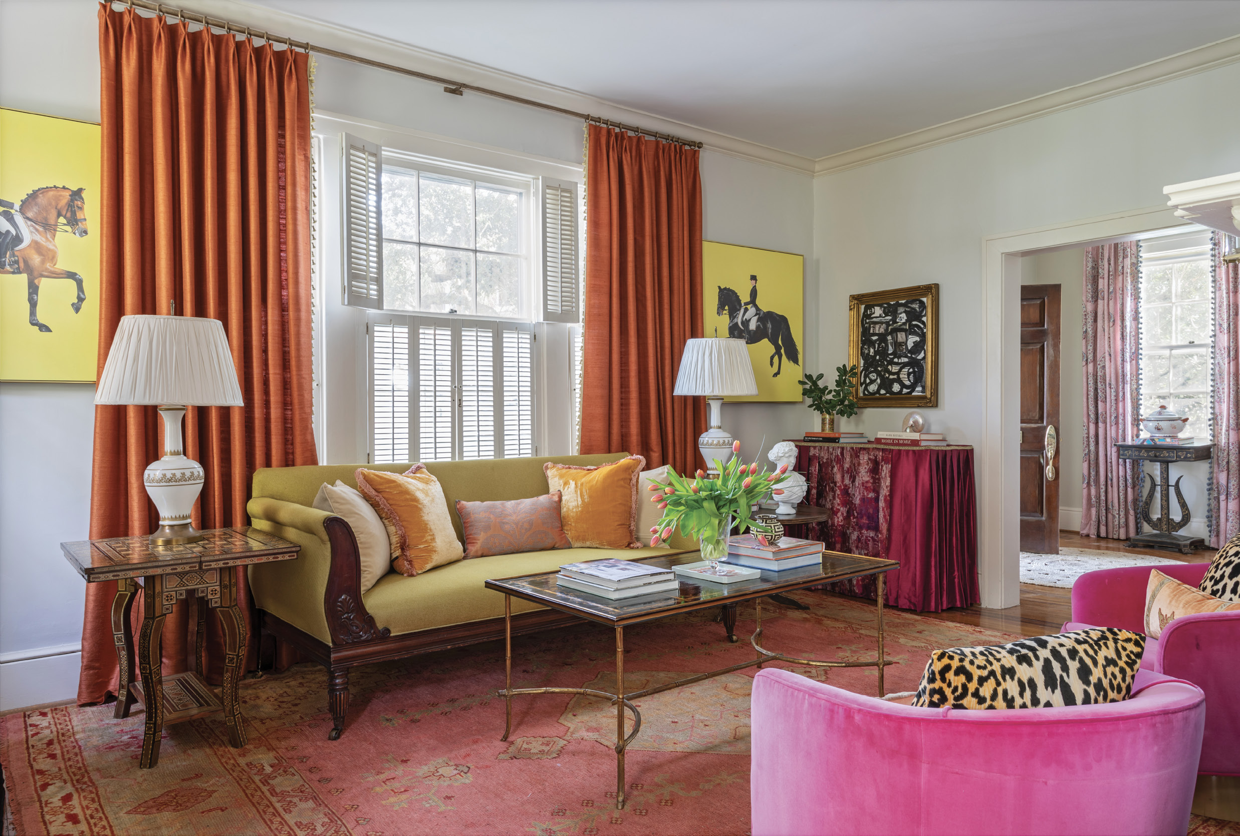 TechniColor Dreams: The sumptuous living room sets the tone for the house. North facing, it can be dark, so Ralph mixed shades of pinks, oranges, and a splash of chartreuse to give it a bright, energetic feel. Brilliant yellow equestrian art by Scout Design Studio and a velvet table skirt pop against the walls painted in Benjamin Moore’s “Pale Oak,” and a vintage coffee table complements the antique sofa upholstered in a woven Sabina Fay Braxton fabric.