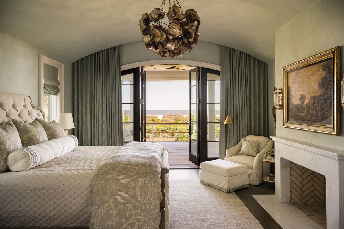 The primary bedroom features a curved plaster ceiling and walls, enhancing the warmth of the cozy space. Susan selected an oil painting of the English countryside from her Gainesville home for above the fireplace, opting to hide the TV in a nearby armoire. “I love that piece. I’d rather look at it than a TV,” she says. Deep green drapes and patterned bedding from Lewis &amp; Wood, Imogen Heath, and Cowtan &amp; Tout add interest.