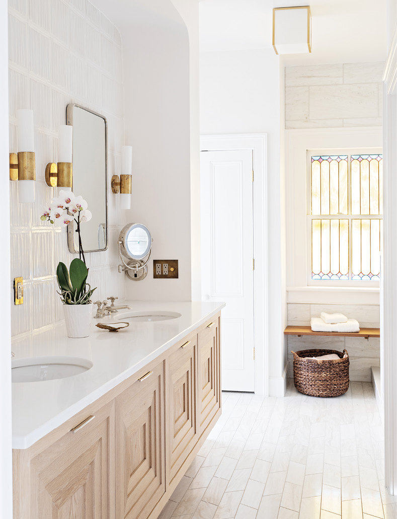 A hint of color from the original stained glass window is picked up in the brass wall sconces by Aerin. The white oak vanity with a stepped-front detail floats in the opposing arch from the adjoining bedroom.