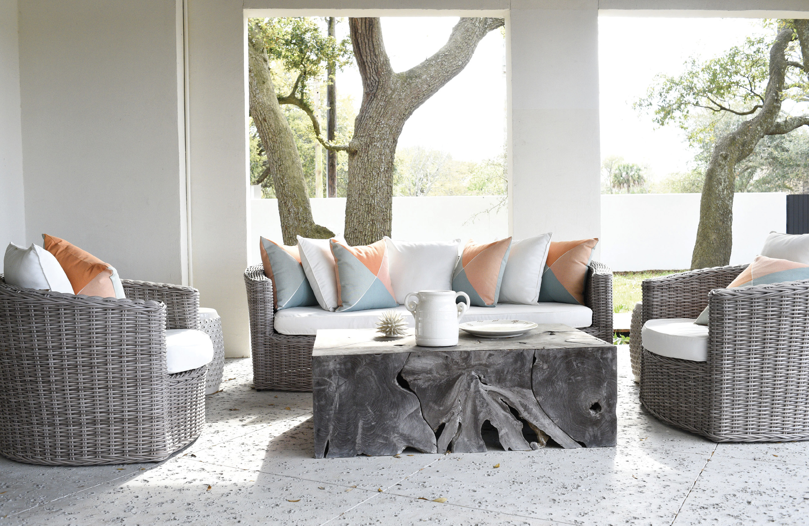 Grey wicker furniture from Celadon Home anchors the outdoor lounging space, with a pop of color provided by Serena and Lily pillows; the spectacular Artesia coffee table from GDC Home pays homage to its deep-rooted neighbors