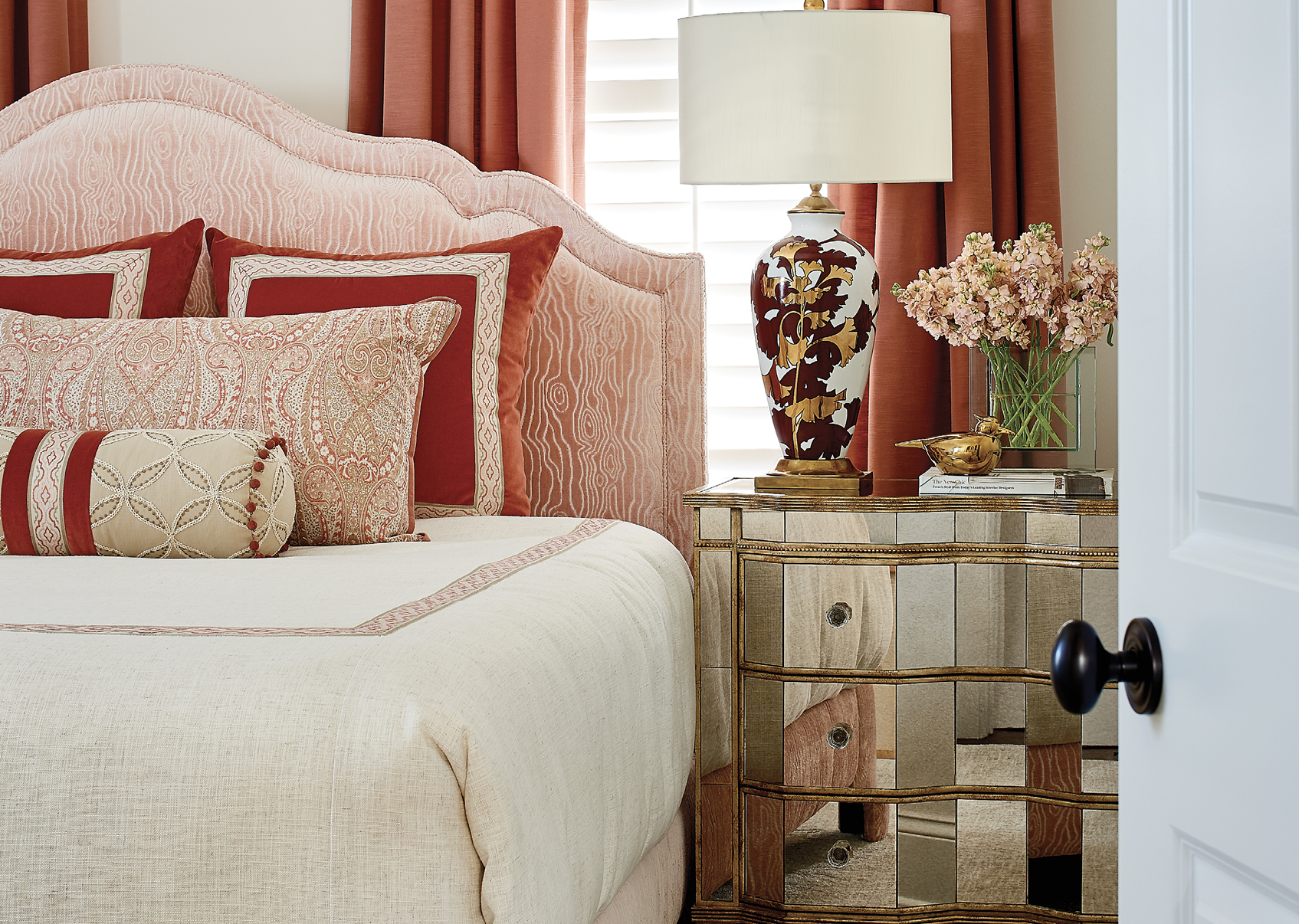 In contrast, the guest bedroom is a cacophony of cheerful colors, with deep peach drapes, a mirrored antique nightstand, and an elegant gold and crimson lamp from Currey and Company.
