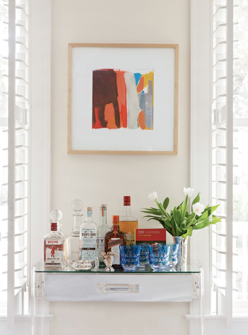 Create Space - A petite Lucite desk with a glass top is the perfect fit for an unobtrusive serving station. The colorful painting by Sally King Benedict helps anchor the mini bar on a neutral wall.  Location: I’On, Mount Pleasant (owned by Allison and LeGrand Elebash)  Issue: March 2016, “Outside the Box”  Photographer: Julia Lynn