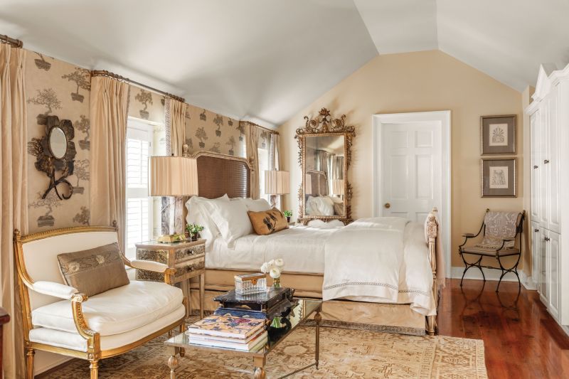 The Suite Life: The primary bedroom occupies the top floor of the converted kitchen house, its gable roof creating a cozy feel in the warm space, painted in Benjamin Moore “Monroe Bisque.” The Jim Thompson “Cedros” wallpaper, featuring metallic bonsai trees, creates an accent wall behind the bed. The motif is replicated with antique textiles crafted into pillows by Cynthia Murphy Designs. The circa-1780 Italian neoclassical gilt-wood mirror from Navona Antiques and Persian Heriz rug from Lavender Oriental Carpets add majestic touches to the space.