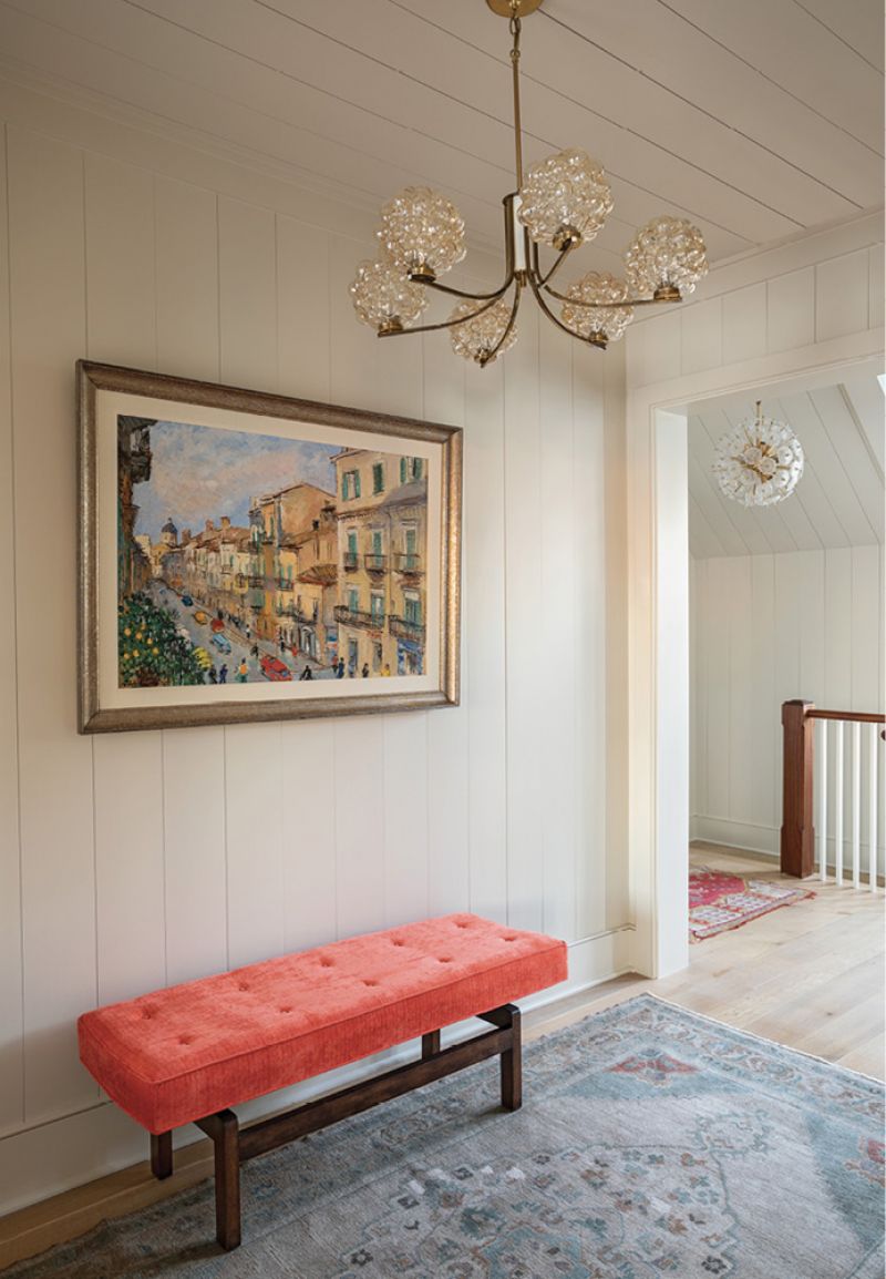 In the upstairs hallway, a wooden bench, upholstered in a playful coral fabric, picks up on hues in the painting of an Italian streetscape and complements the whimsical Val Saint Lambert Sputnik chandelier.