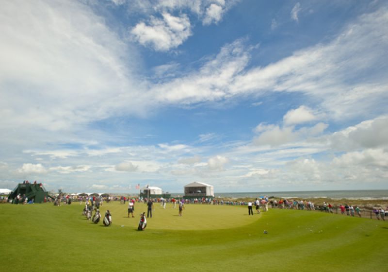The putting green loaded with pros under a clearing sky.