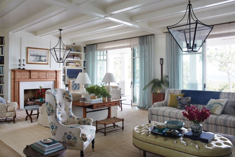 The light, bright interiors painted in “James White” by Farrow &amp; Ball pull in hues from the picturesque view, including pops of green and blue and light-colored woods. A large sisal rug from Eve and Staron Studio anchors the space with comfortable furniture in pretty patterns and a green leather ottoman/coffee table by Nietert Antique Restoration.
