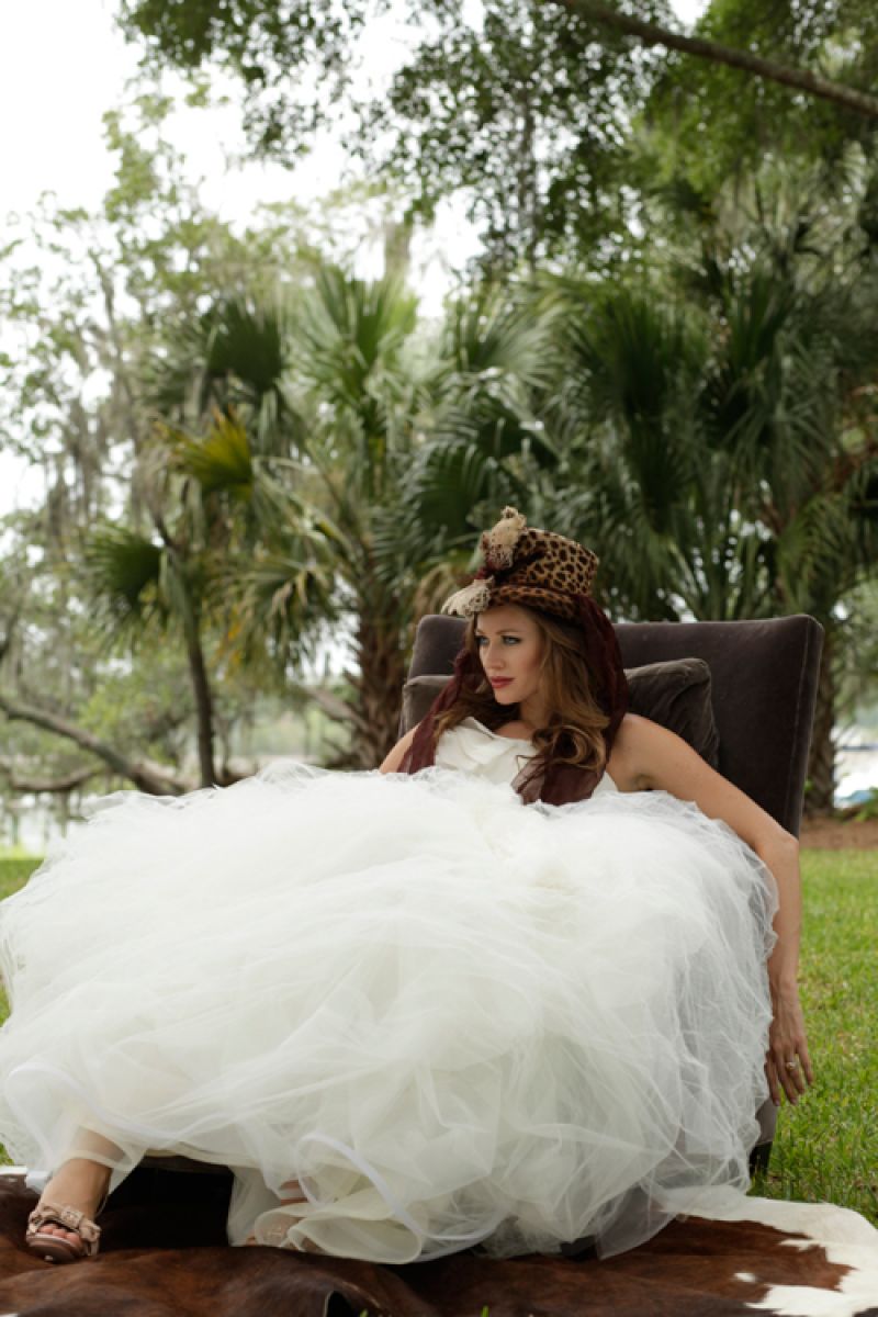 Vera Wang’s “Jocelyn” gown with distressed organza, tulle, and lace tiered skirt from Maddison Row. Jade, chalcedony, and freshwater pearl flower pendant necklace from Theodosia. Badgley Mischka’s satin, suede, and leather heels with crystal embellished bow from Gwynn’s of Mt. Pleasant. “Liza” felt hat with pheasant feathers and organza from Magar Hatworks. Chair and cowhide rug from RiverOaks personal collection.