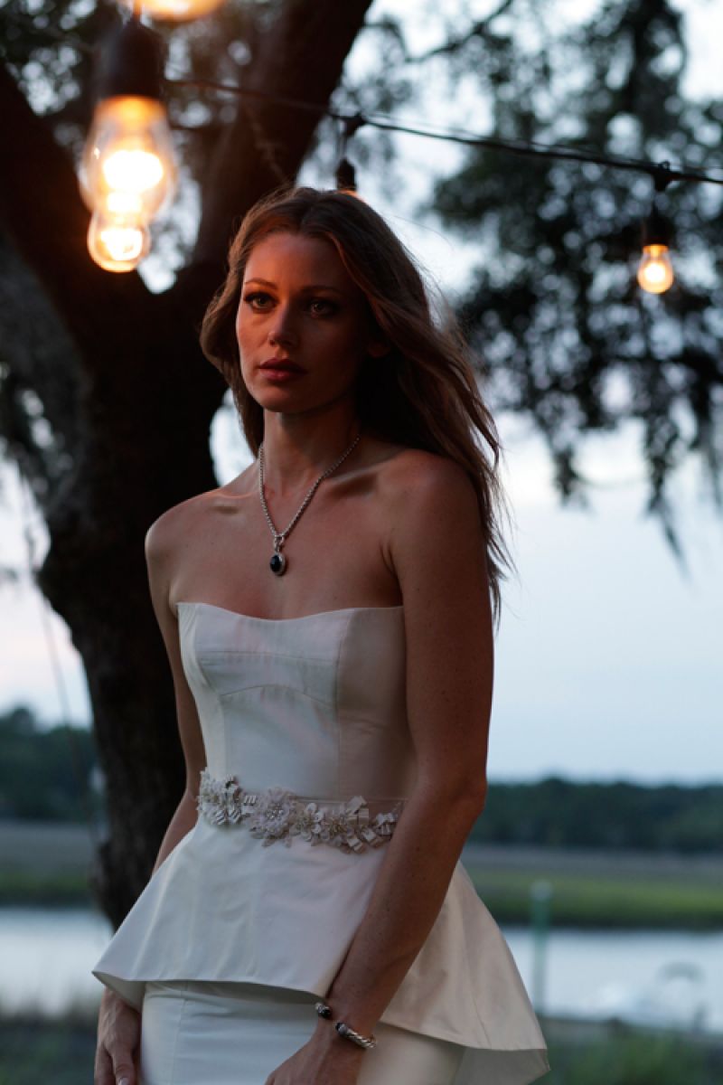 Legends by Romona Keveza gown with crystal embellished floral belt and detachable peplum from Maddison Row. David Yurman’s sterling silver wheat chain necklace with onyx and diamond pendant from the Cerise collection and his silver, hematite, and diamond bracelet from his Moonlight Ice collection, both at REEDS Jewelers. Rhinestone bow hair clip from Jean’s Bridal.