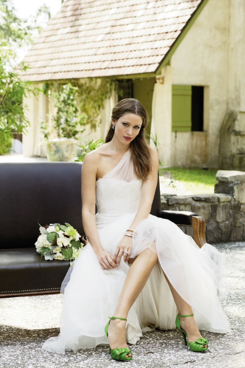 Monique Lhuillier’s “Charlene” gown with one-shoulder illusion neckline  and tulle skirt from Maddison Row.  Three 14K yellow gold and silver cuff bracelets with  diamond accents from Joint Venture Estate Jewelers.  Platinum and diamond earrings and 14K white gold ring with diamonds, both from Croghan’s Jewel Box. Nina’s d’Orsay heels from LulaKate. Bouquet by Jennigray Hewitt of RiverOaks. Sofa from Muster Branch Vintage Rental Co. by Lauren Heuber.